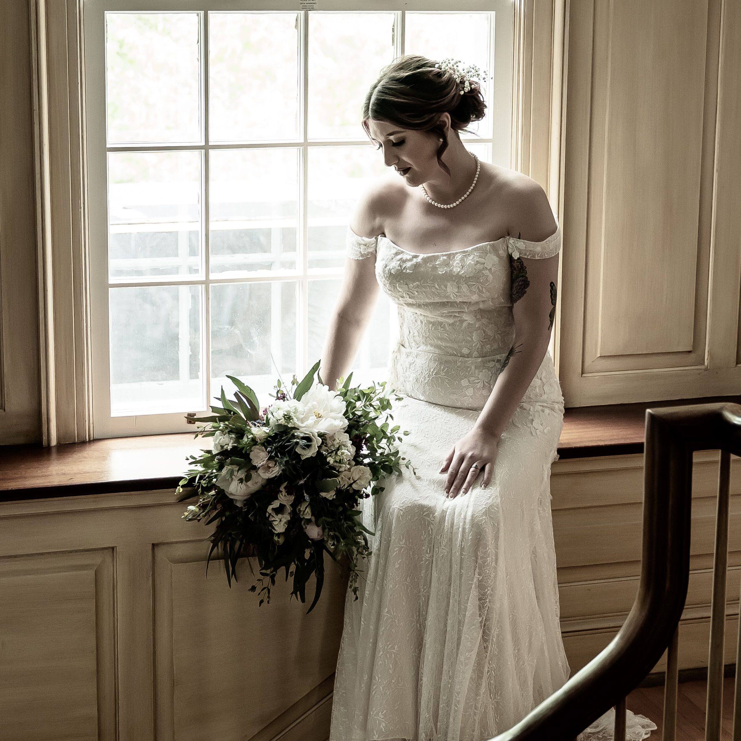 A bride sits with her bouquet in front of a large window, the light charming her white gown. She looks down at the flowers at her side, a hand resting on her knee.